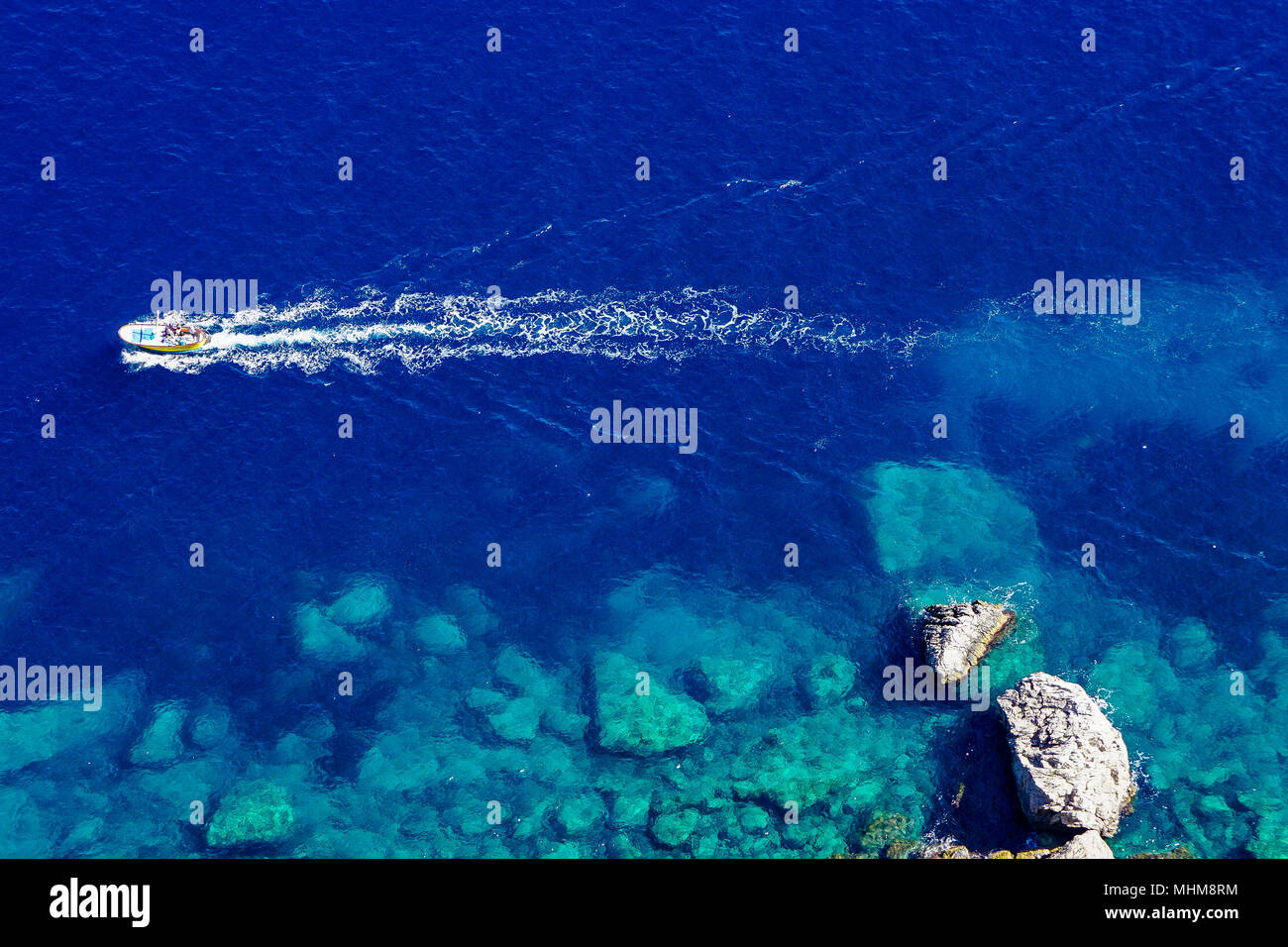 Traditional gozzo boat of Capri, Italy in crystal clear water with trailing wake shot from cliffs far above Stock Photo