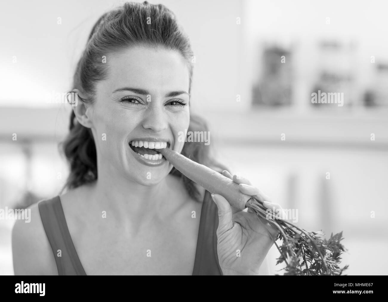 Happy young woman eating carrot in kitchen Stock Photo