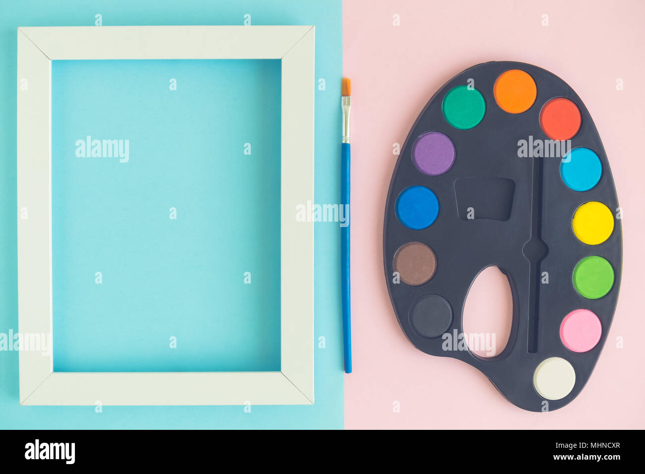 Art palette with paints, photo frame and paintbrush against pastel pink and blue background Stock Photo