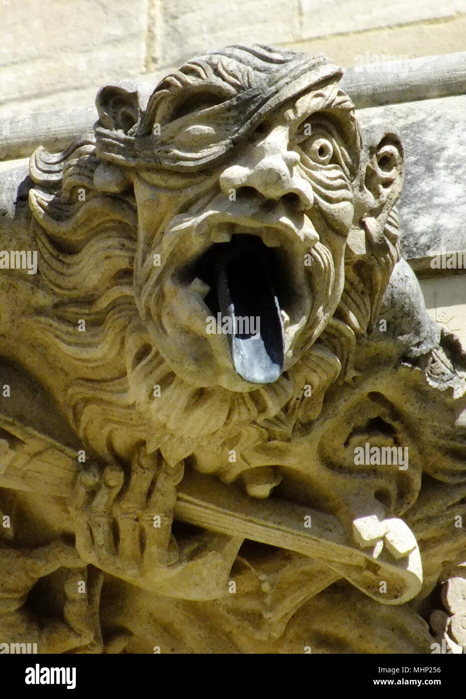 Gargoyle of an ugly man playing a stringed instrument, with a water spout in his mouth, at Gloucester Cathedral, Gloucestershire. Stock Photo