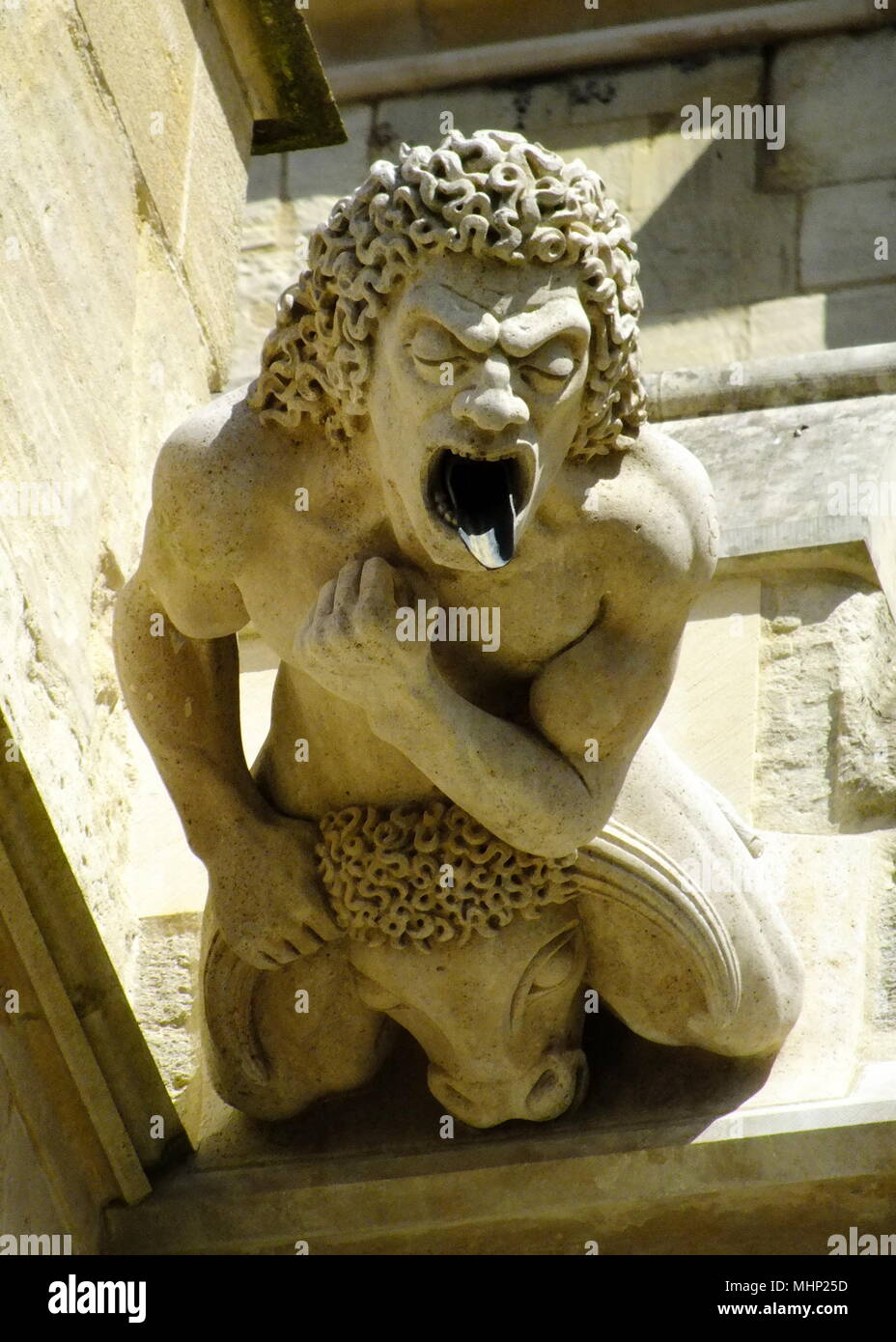 Gargoyle of an ugly man riding on a bull, with a water spout in his mouth, at Gloucester Cathedral, Gloucestershire. Stock Photo