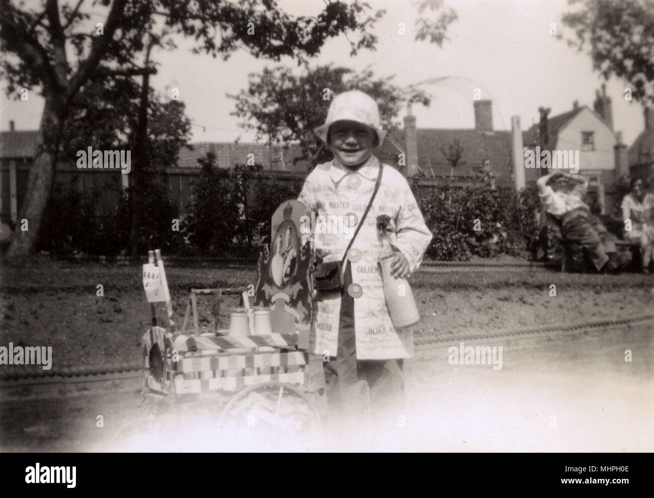 A little girl dressed up as a milkman (or milk woman), complete with her miniature milk float.  The photograph of King George VI and Queen Elizabeth probably dates this image as 1937 as it seems a likely costume to be included in a carnival or parade to celebrate the Coronation that year.       Date: 1937 Stock Photo