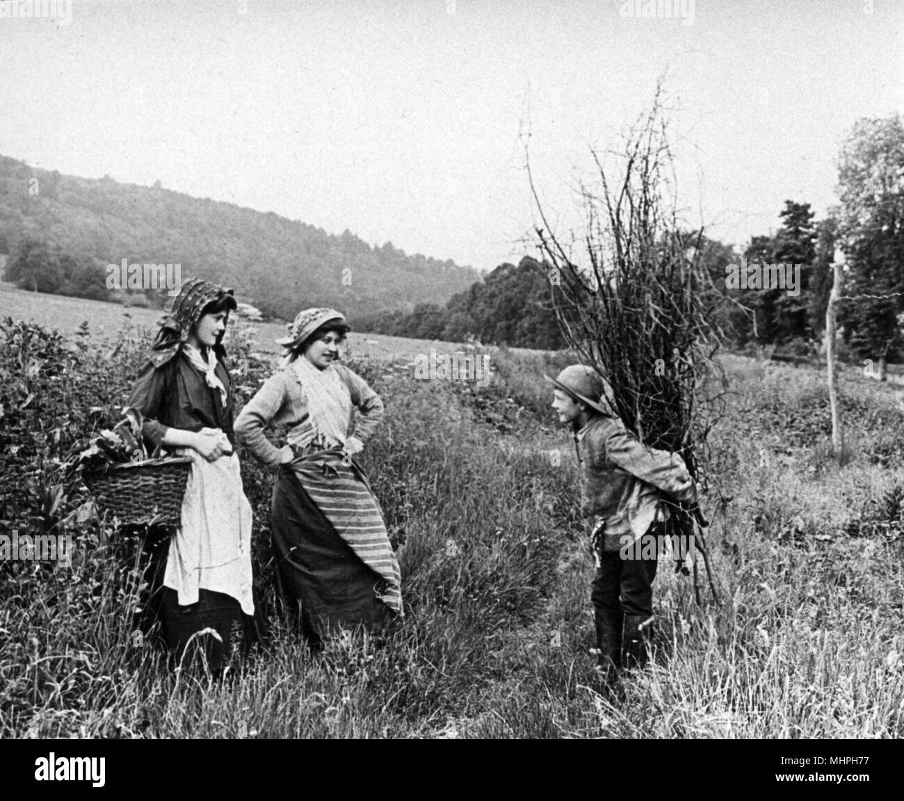 Two young countrywomen greet a boy who has been collecting a bundle of sticks, 1890s     Date: 1890s Stock Photo