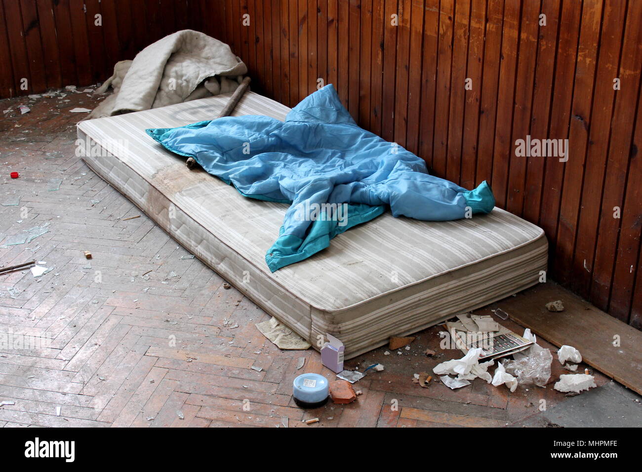 Abandoned room with old mattress, cover, stick, hand lotion, open condom wrappers, tissues, old newspaper, broken glass and garbage used by homeless a Stock Photo