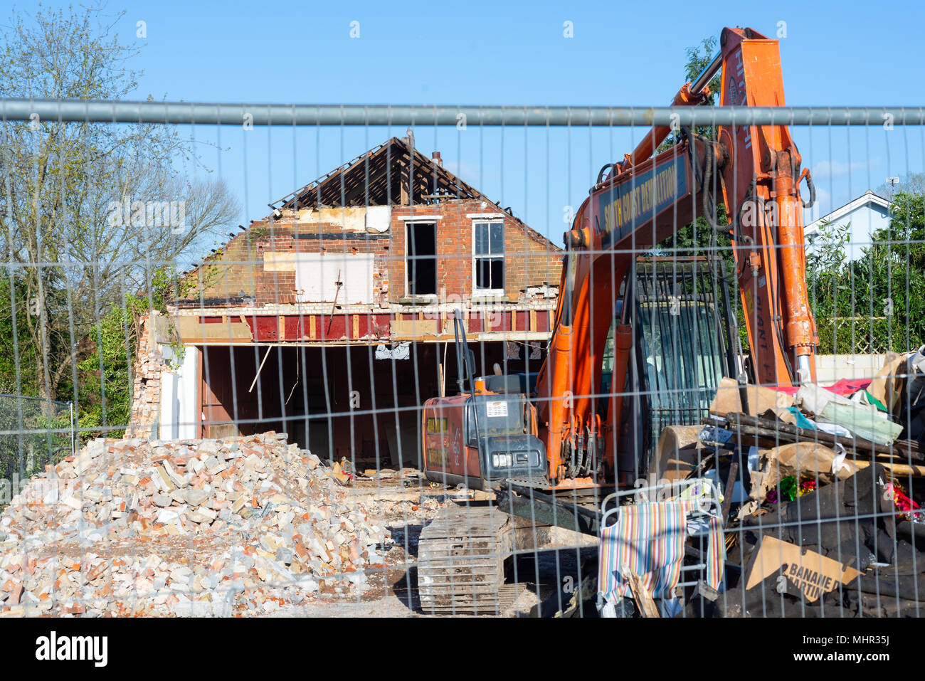 Demolition site with a long arm digger, piles of rubbish and rubble, surrounded by a metal security fence, UK Stock Photo
