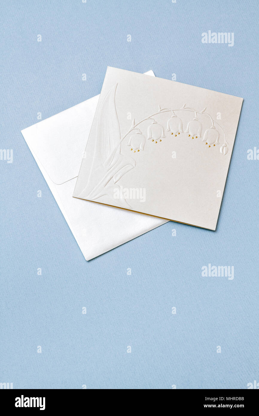 White wedding invitation with Lily of the valley flowers emboss on light blue paper background. Vertical shot. Shiny pearlescent card with envelope. Stock Photo
