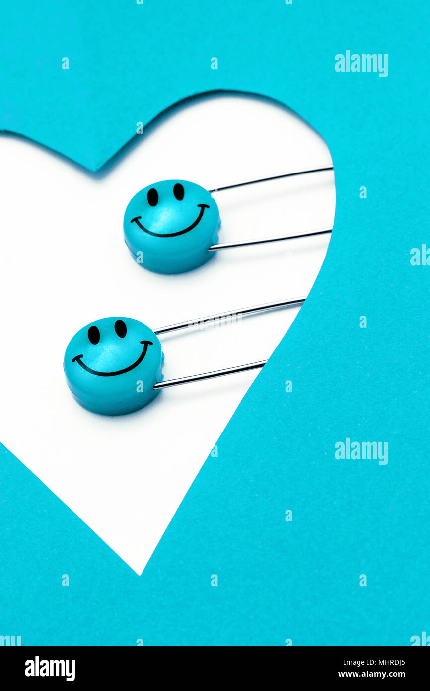 Plastic head metal safety pins pinned in heart shaped paper. Blue cyan smile emoticon safety pin. Cute and funny colorful emoticons. Stock Photo