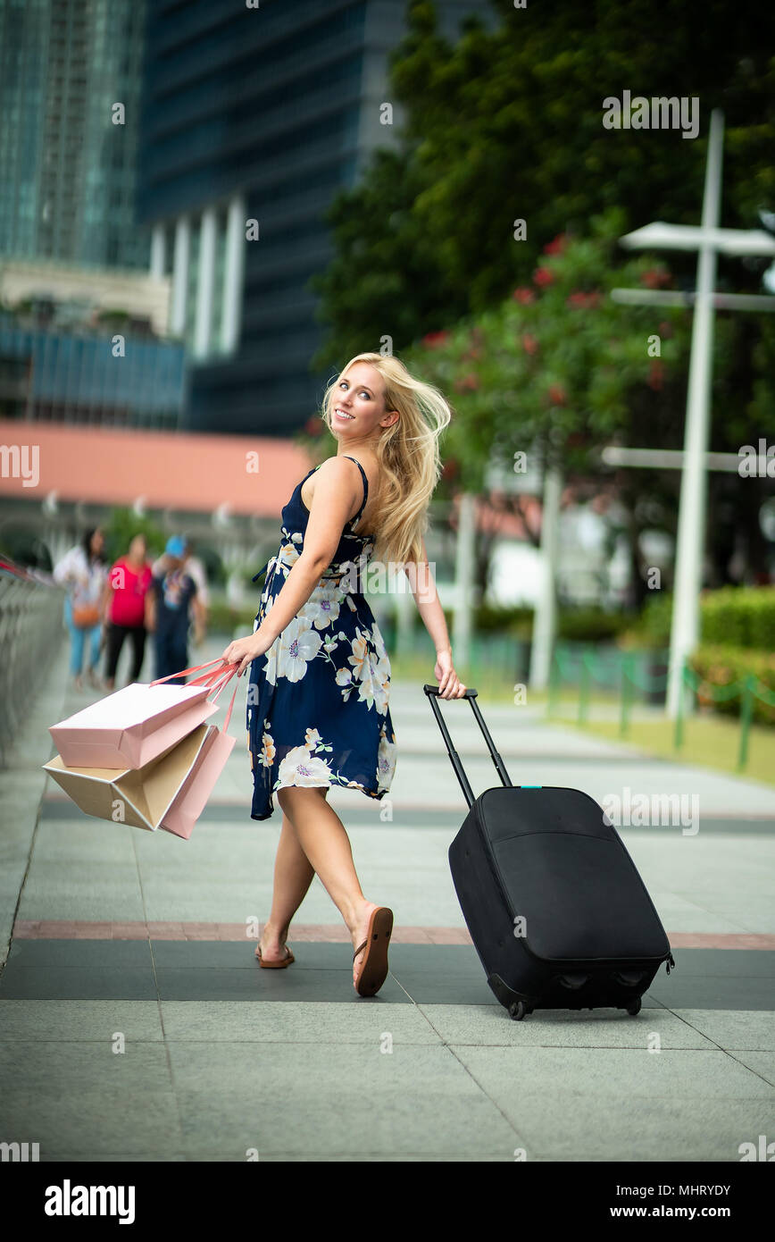 Happy Traveler after shopping. Excited Beautiful Woman wearing casual blue dress with flowers holding Black suitcase and shopping bags in the stree Stock Photo Alamy