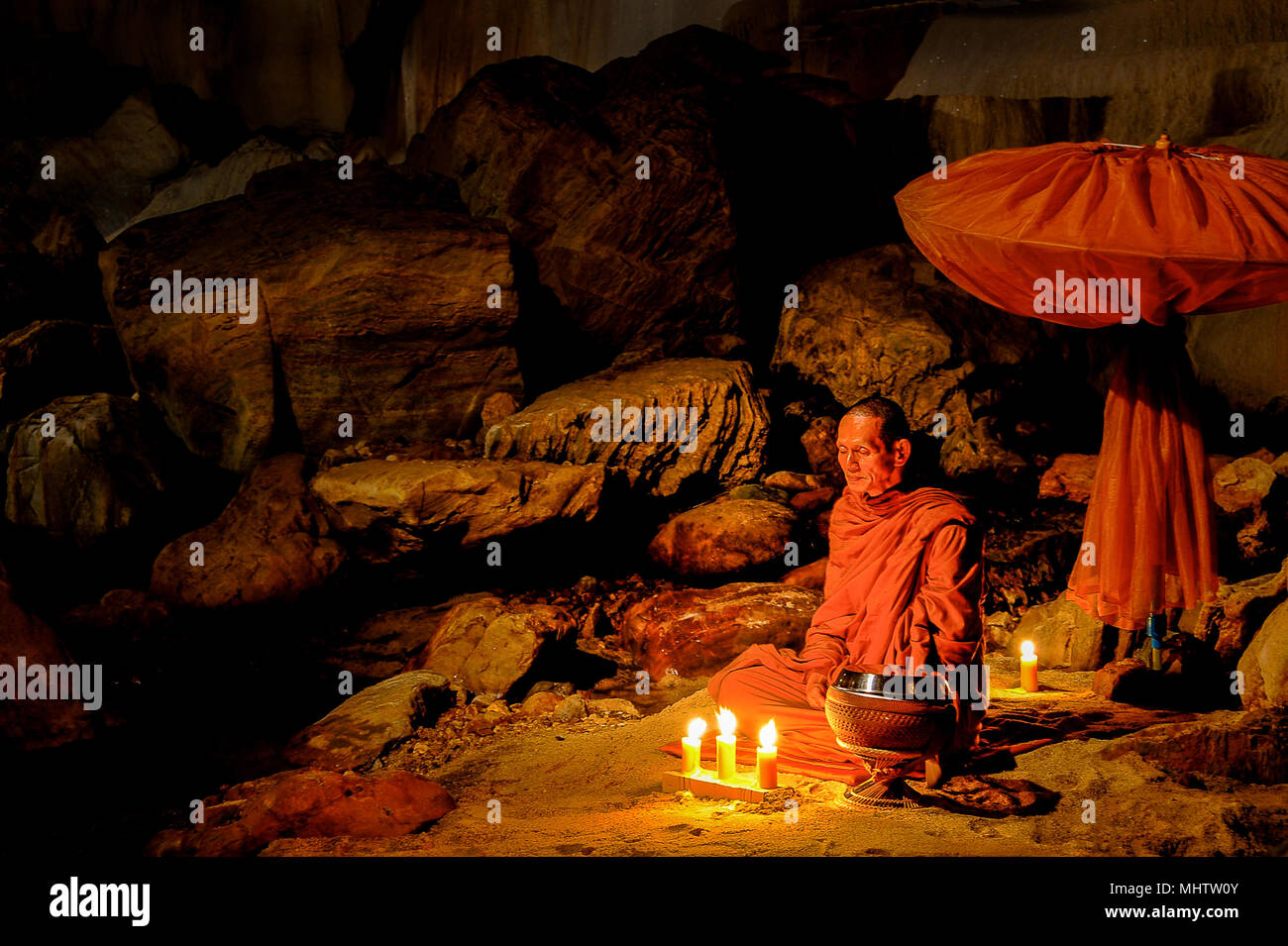 Kanchanaburi, Thailand - July 15, 2011: Buddhist monk sitting with alms bowl, lighten candle and long-handles umbrella to do individual meditation in  Stock Photo
