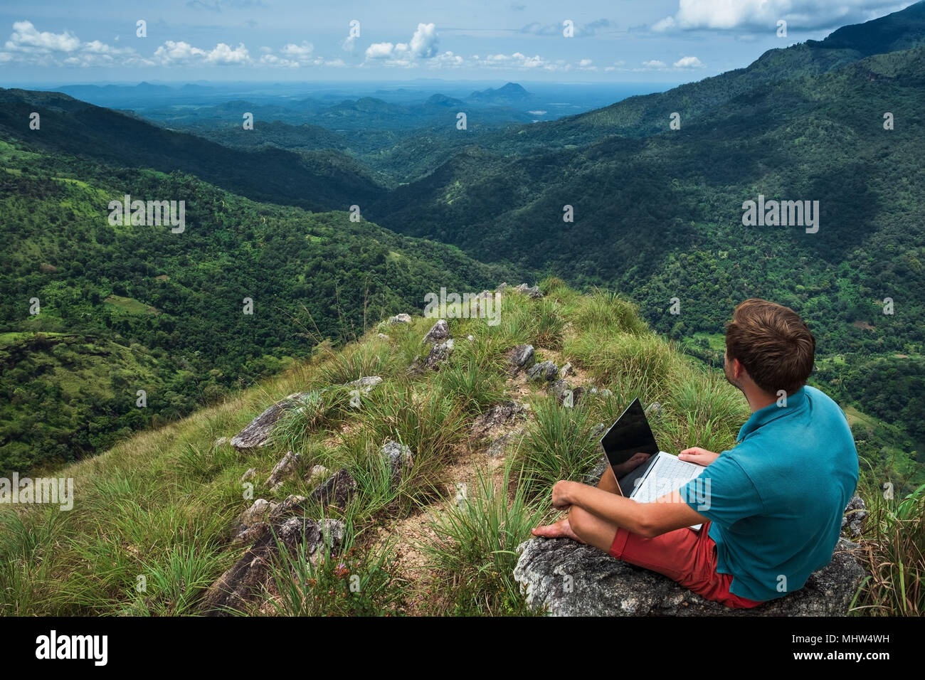 caucasian man with laptop sitting on the edge of ella mountain with stunning views of the valley in Sri Lanka. Stock Photo