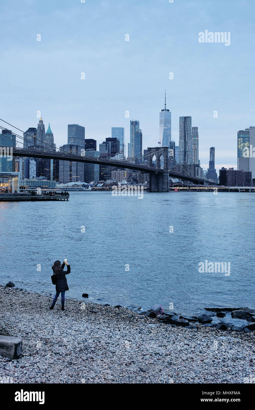 Woman photographing Brooklyn Bridge and Lower Manhattan Skyline from riverbank, New York, USA Stock Photo