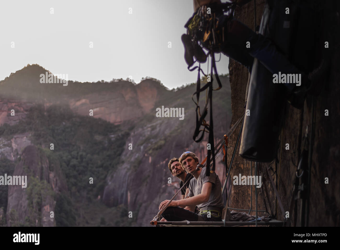 Two rock climbers sitting on portaledge, watching friend climb rock beside them, Liming, Yunnan Province, China Stock Photo