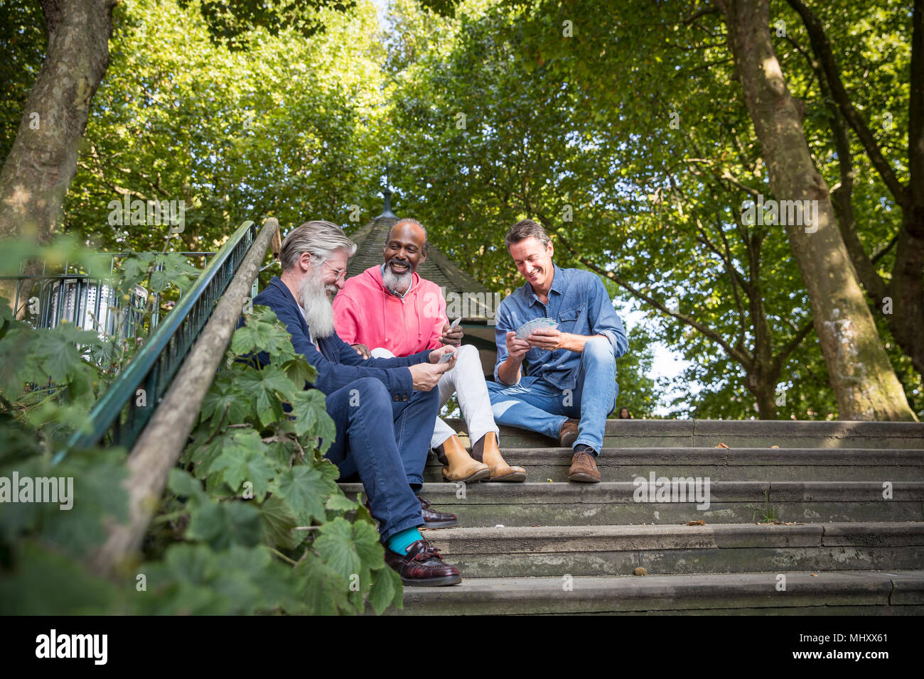Three mature men, outdoors, sitting on steps, playing cards, low angle view Stock Photo