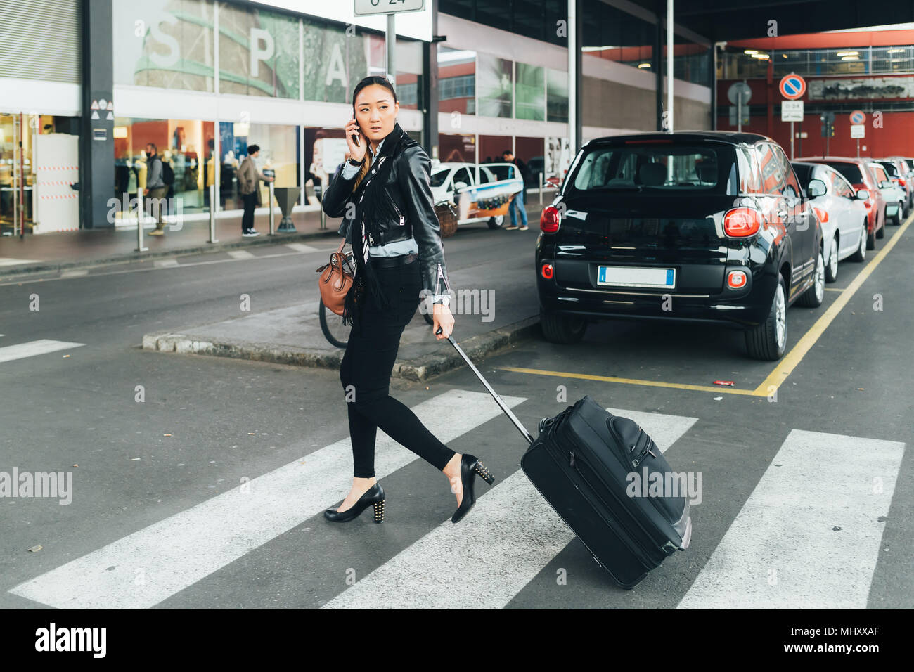 Woman walking outdoors, pulling wheeled suitcase, talking on smartphone Stock Photo