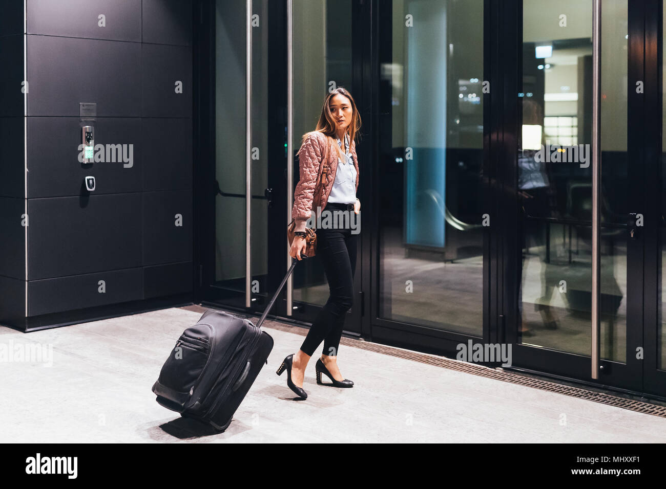 Woman walking towards hotel, pulling wheeled suitcase, looking over shoulder Stock Photo