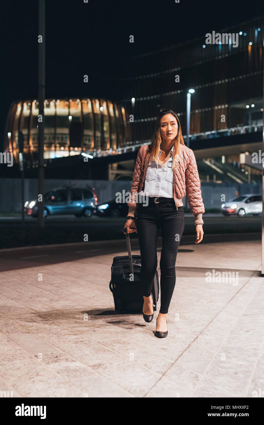 Woman walking outdoors at night, pulling wheeled suitcase Stock Photo