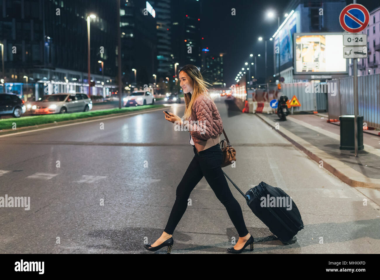 Woman walking outdoors at night, pulling wheeled suitcase, holding smartphone Stock Photo