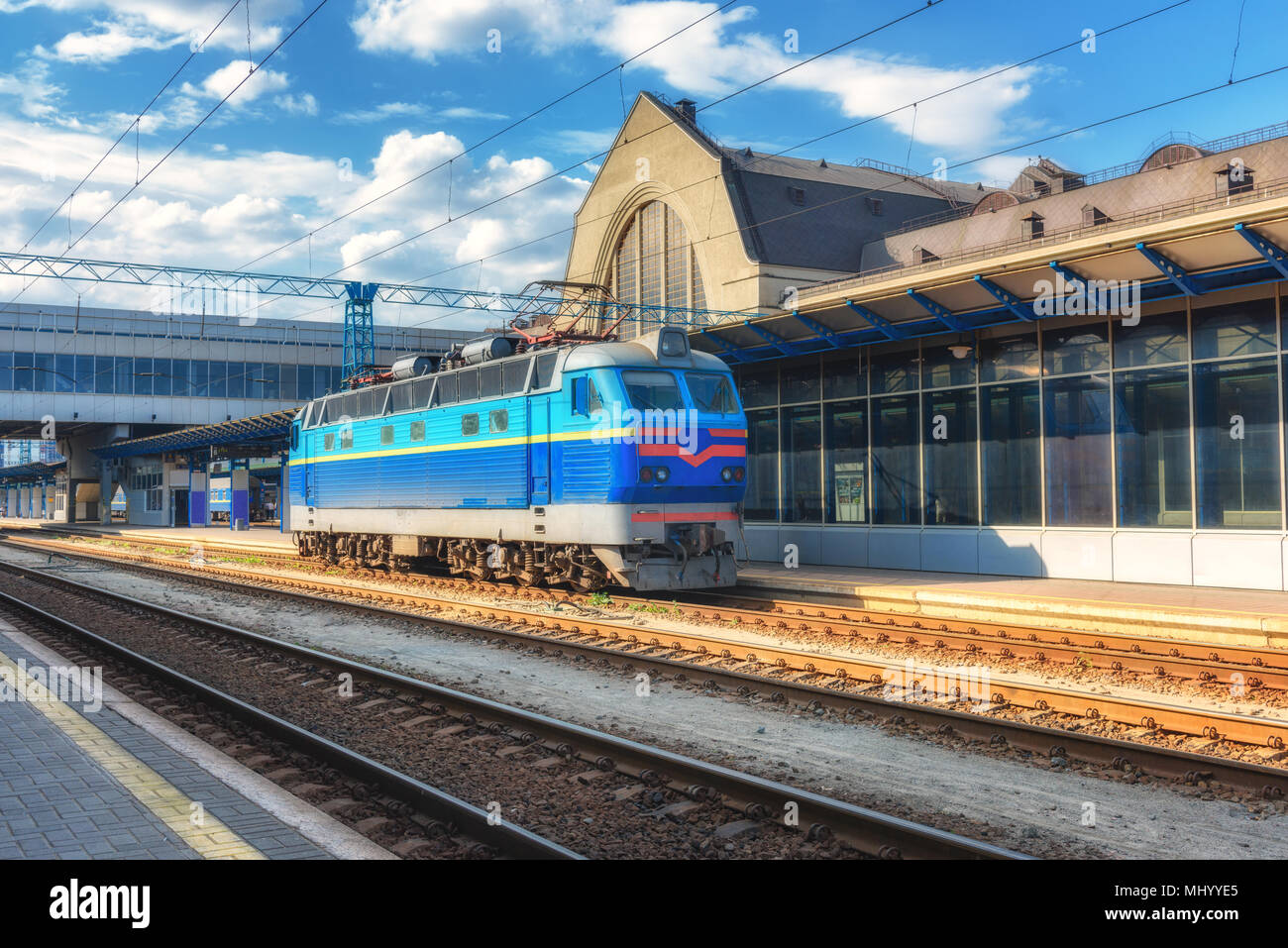 Blue freight train on the railway station at sunset in Ukraine. Industrial landscape with old locomotive, buildings, rails and blue sky with clouds. T Stock Photo