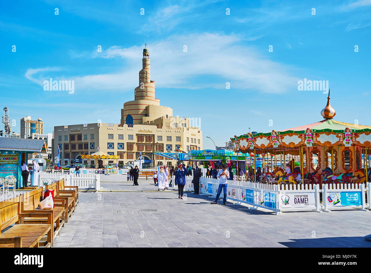 DOHA, QATAR - FEBRUARY 13, 2018: The street of Al Souq district with carousels of Luna park and the scenic building of Al Fanar mosque on the backgrou Stock Photo