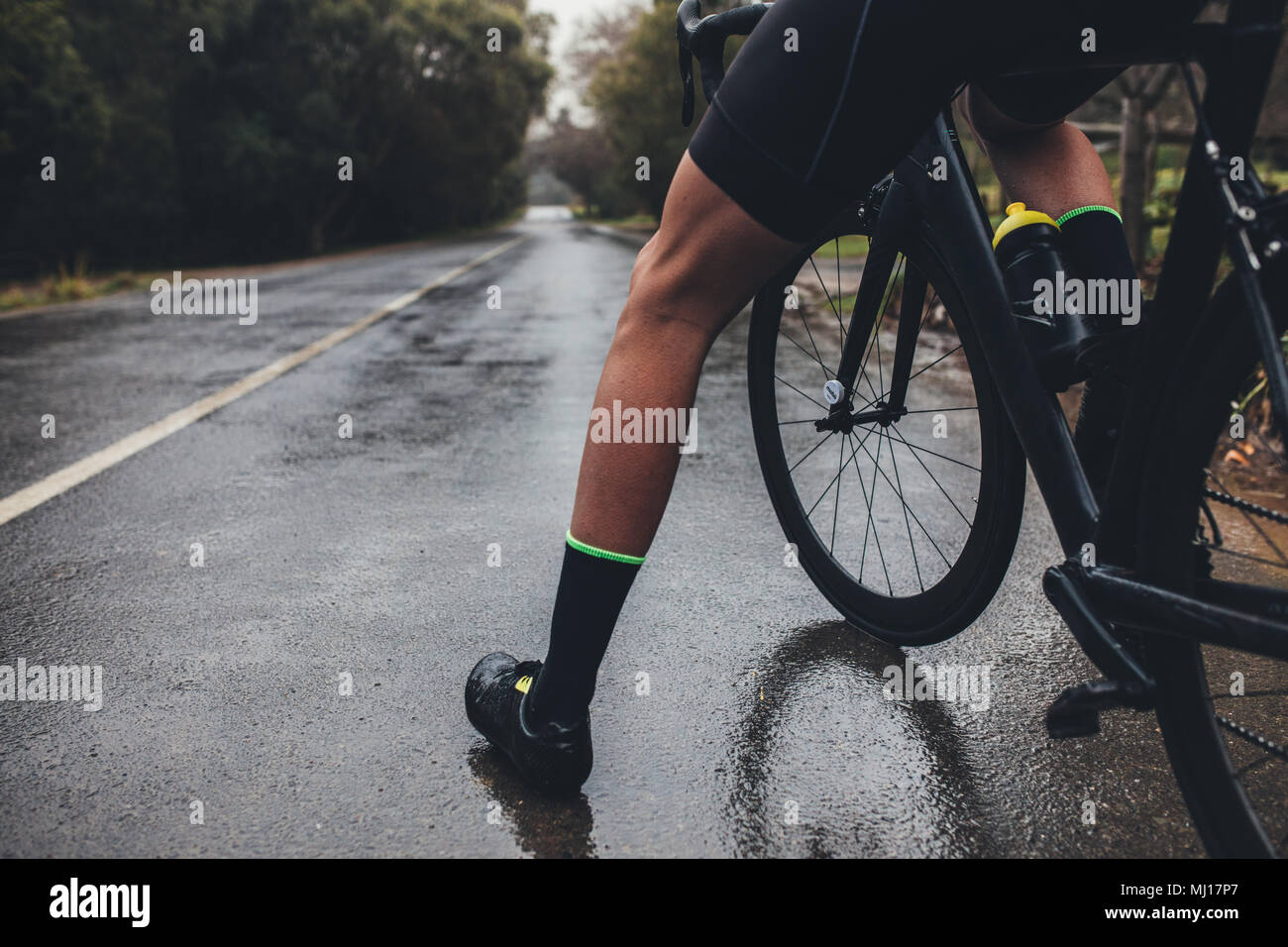 Low section shot of male athlete standing with his bicycle. Man cyclist with his bike on wet country road. Cropped shot of male triathlete with his cy Stock Photo