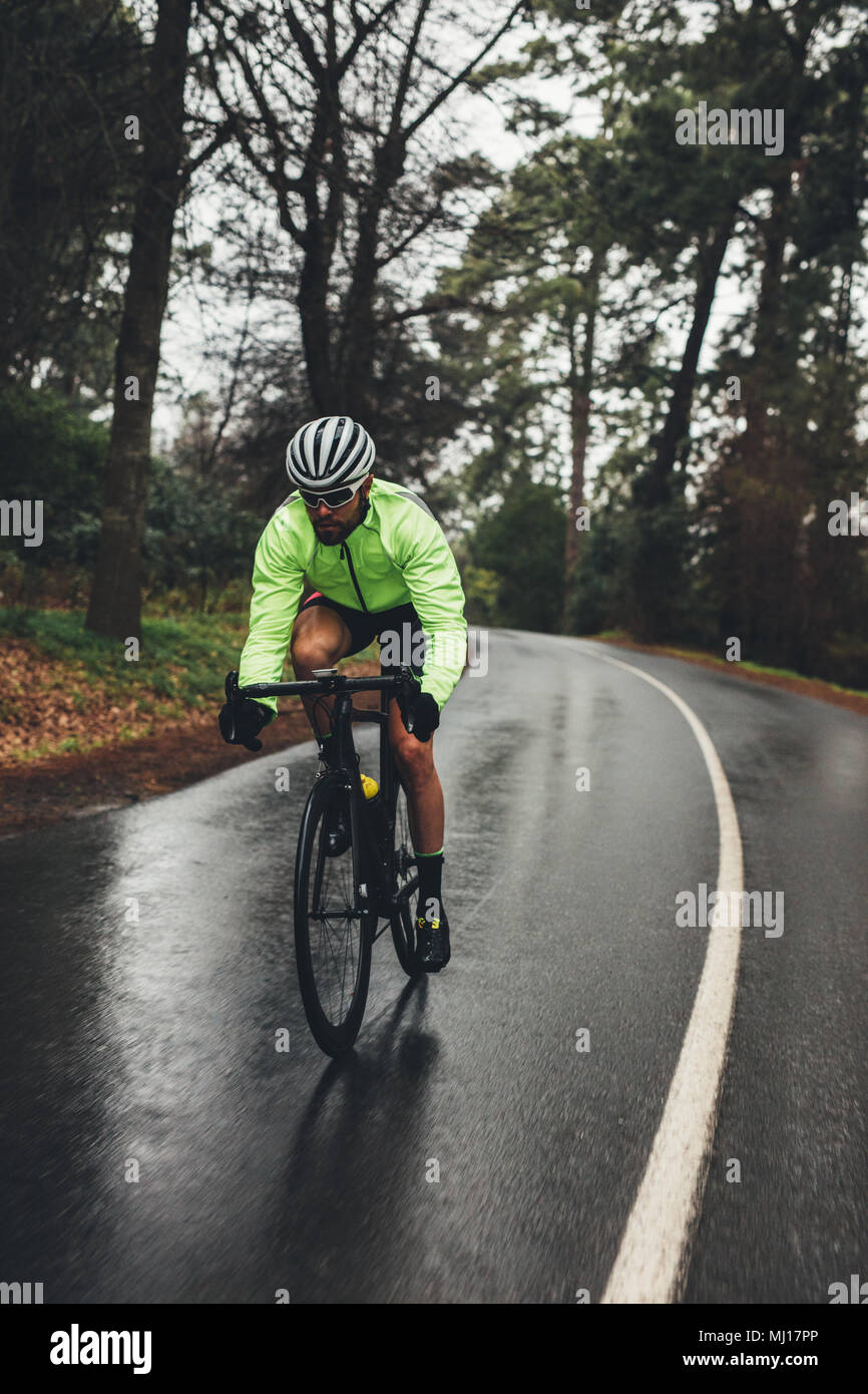 Male cyclist riding bike on countryside highway on a rainy day. Fit sportsman in green jacket cycling on empty road through forest. Stock Photo