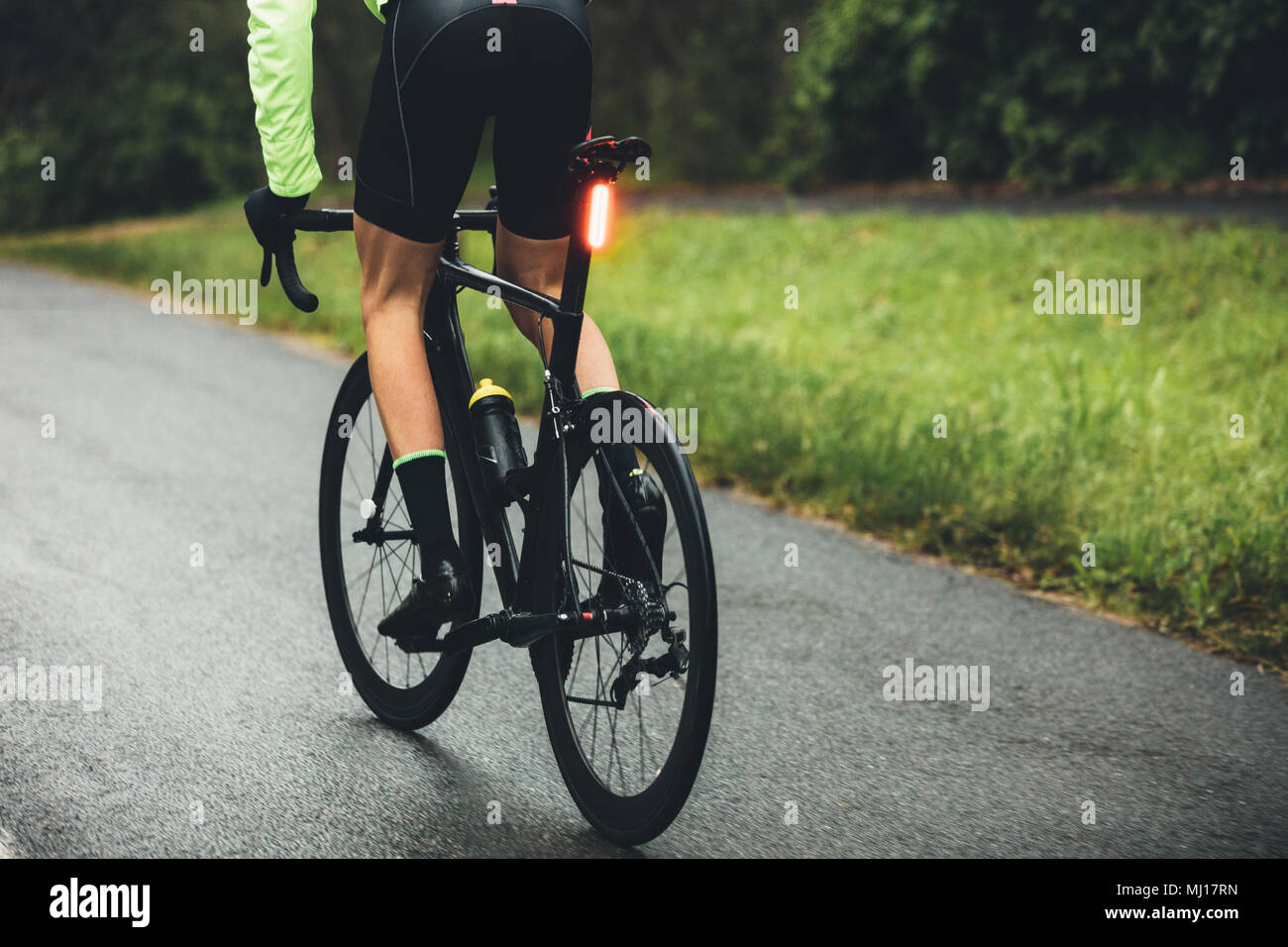 Rear view of male cyclist practising on a wet road in countryside. Professional cyclist riding bicycle on open road. Stock Photo