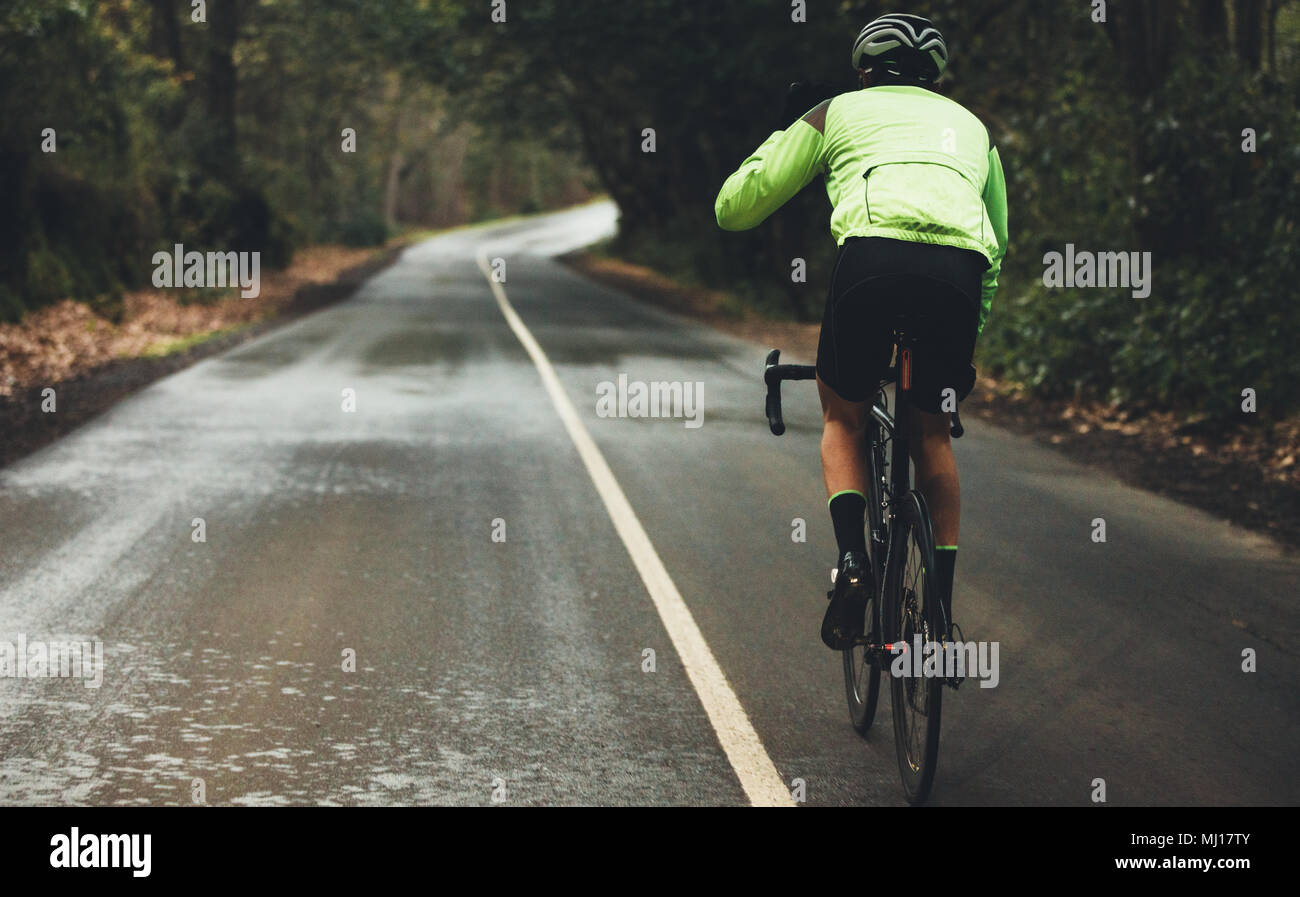 Rear view of male athlete cycling on country road on rainy day. Professional cyclist riding a bike on empty highway through forest. Stock Photo