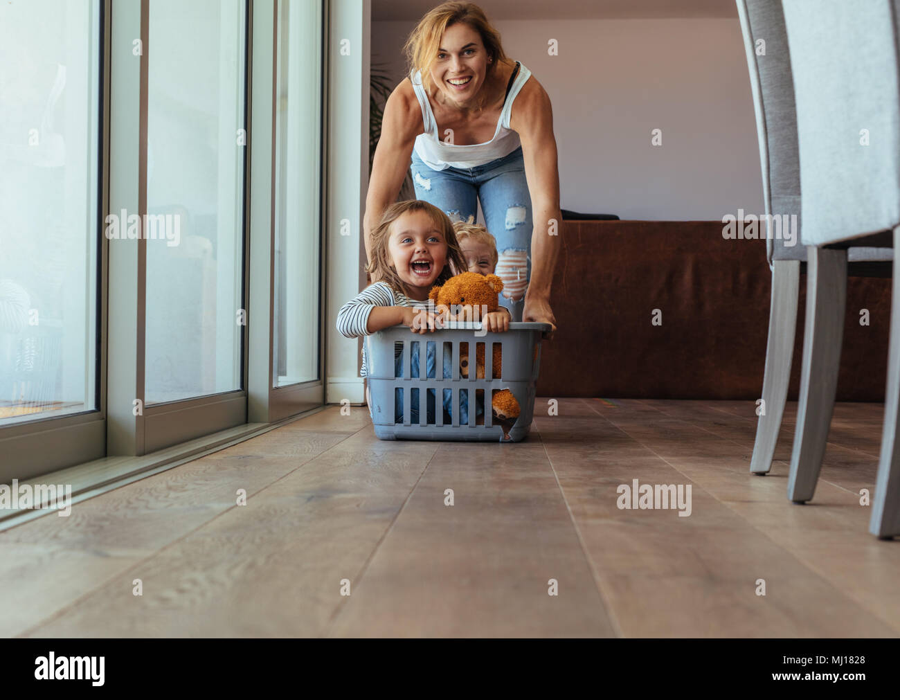 Happy young woman pushing kids sitting in washing basket. Children enjoying laundry basket ride at home. Stock Photo