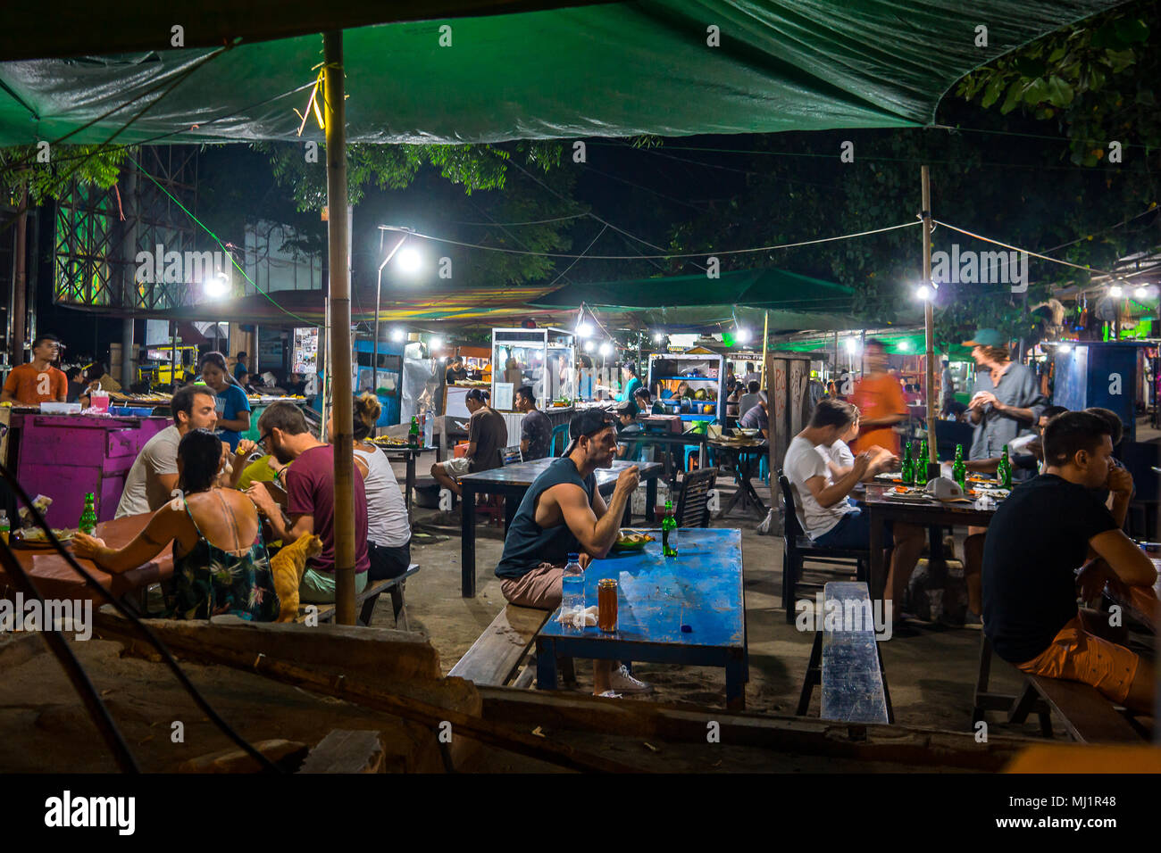 European turists and local residents sitting in the light of electrical bulbs at long tables having various streetfood in an indonesian nightmarket, a Stock Photo