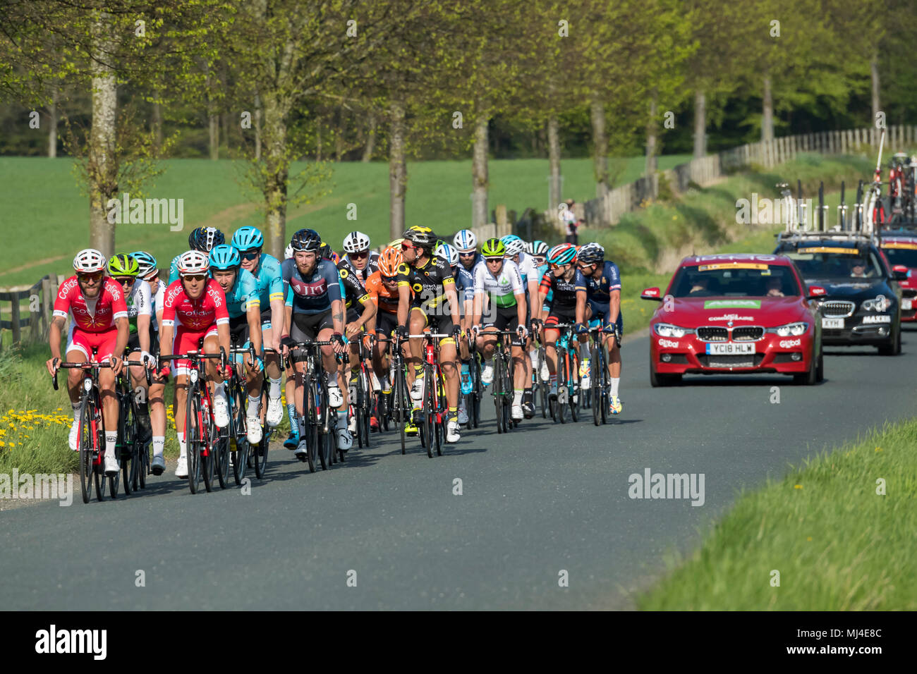 Denton, West Yorkshire, 4th May 2018. Large group of male cyclists in the pelaton, competing in the sunny Tour de Yorkshire 2018, are racing past the grounds of Denton Hall, on a straight, flat, scenic, countryside lane, closely followed by a line of team cars - near Ilkley, North Yorkshire, England, UK. Credit: Ian Lamond/Alamy Live News Stock Photo