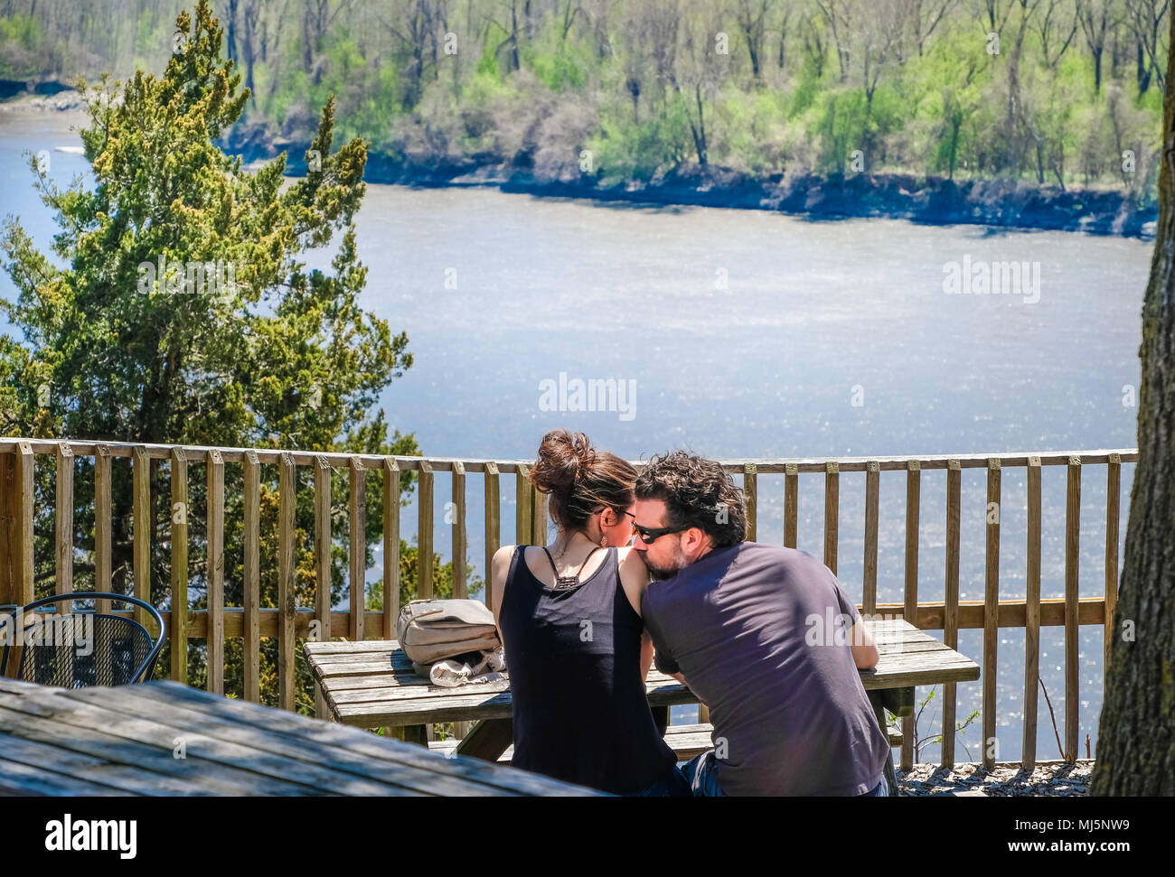 Rocheport, Missouri, USA, April 30, 2018: Young couple sitting by the river; man tenderly kissing the woman's shoulder; river and woods in the backgro Stock Photo