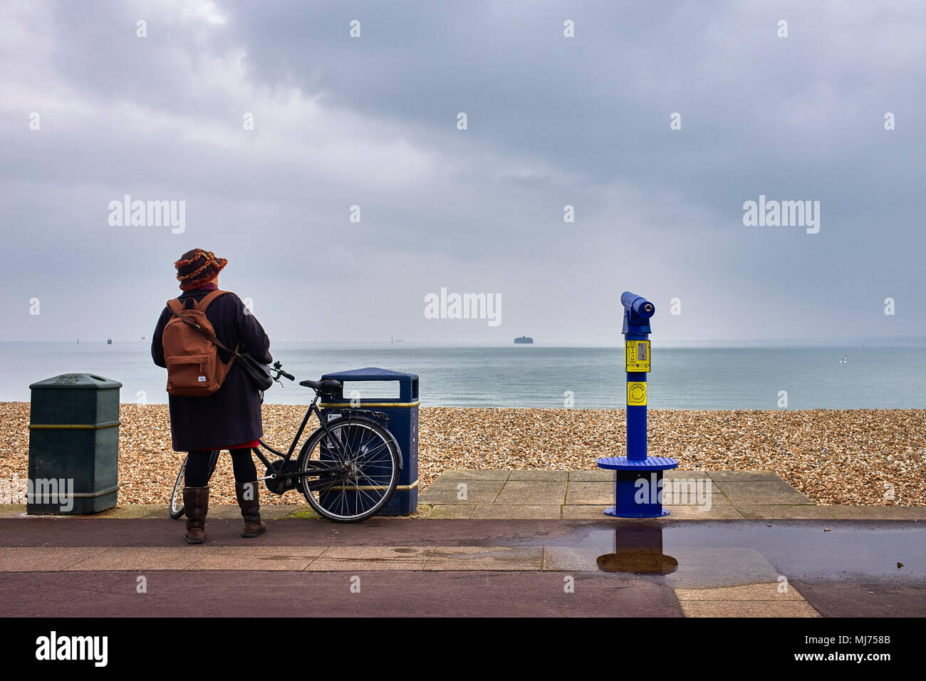 Woman looking out to sea with bicycle next to talking telescope on the promenade at Southsea, Hampshire on an overcast day Stock Photo