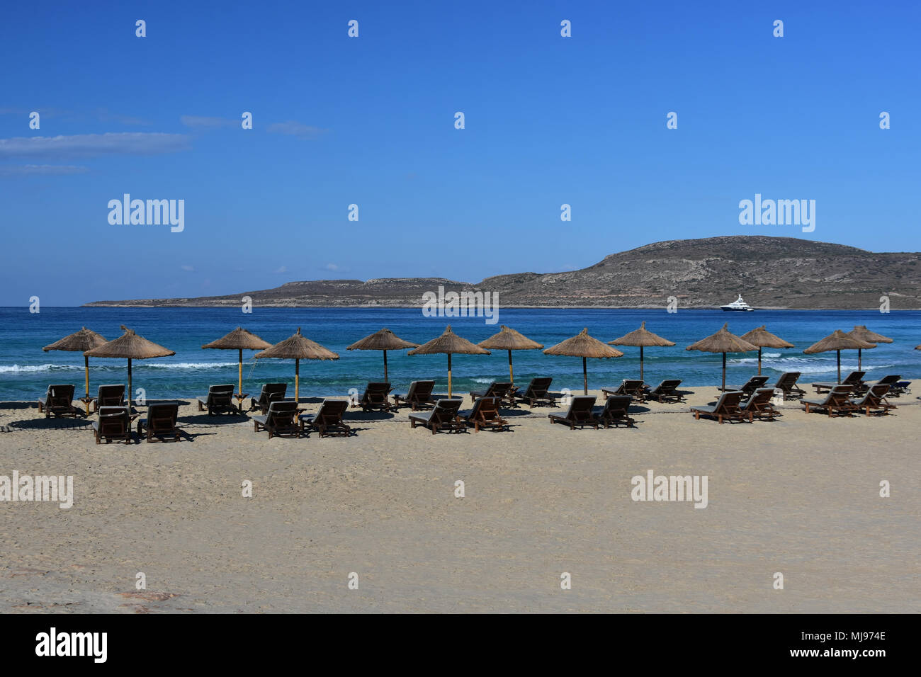 Wide sandy beach with old umbrellas and empty chaise longue, quiet turquoise sea, white yacht far away in the bay, serene blue sky Stock Photo