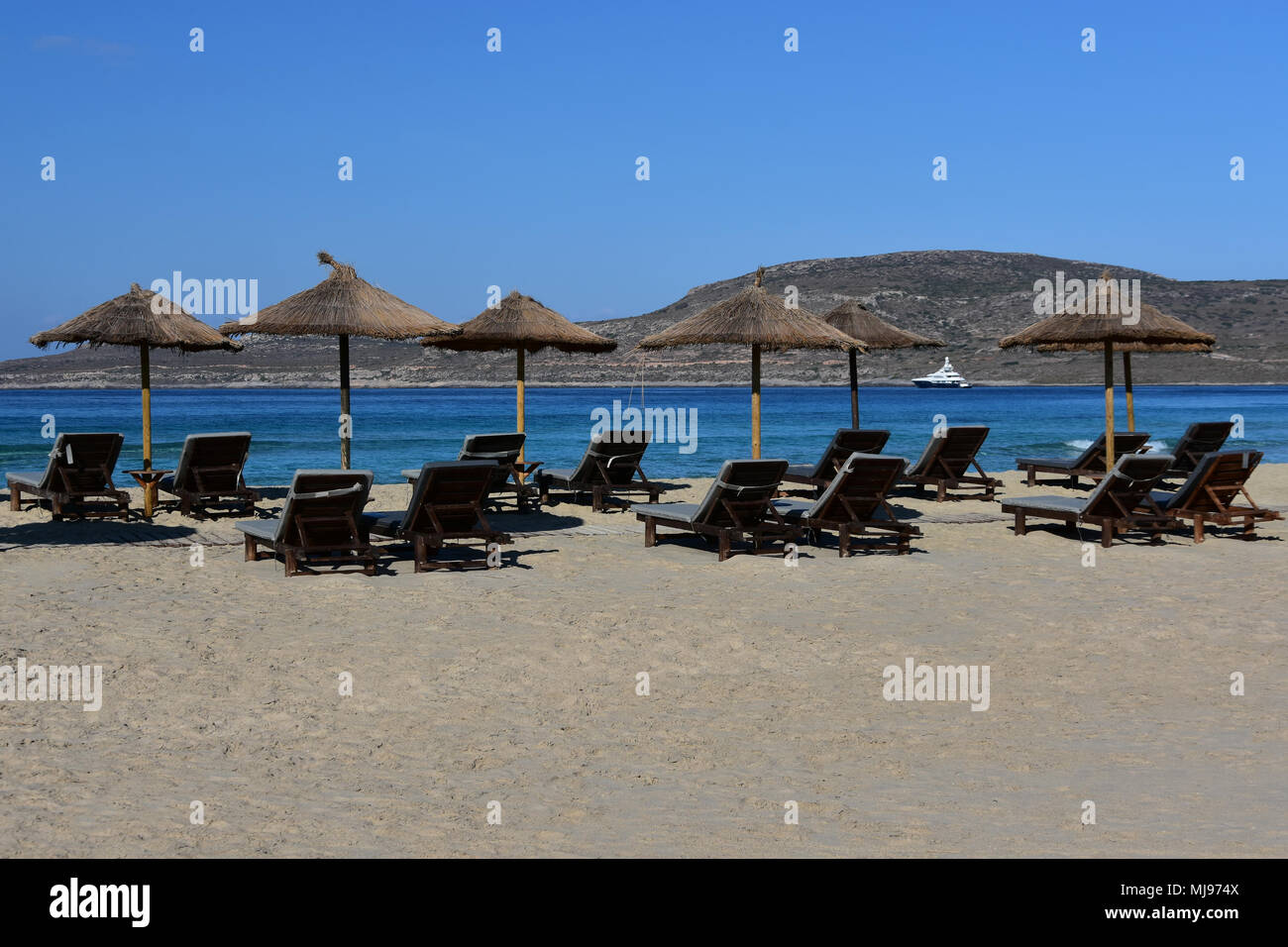 Wide sandy beach with old umbrellas and empty chaise longue, quiet turquoise sea, white yacht far away in the bay, serene blue sky Stock Photo
