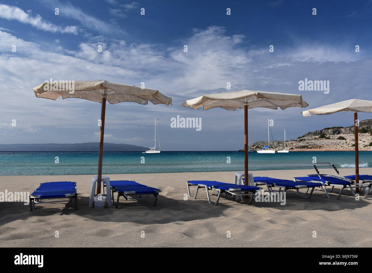Wide sandy beach with old umbrellas and empty chaise longue, quiet turquoise sea, white yachts in the bay, cirrus clouds at sky Stock Photo
