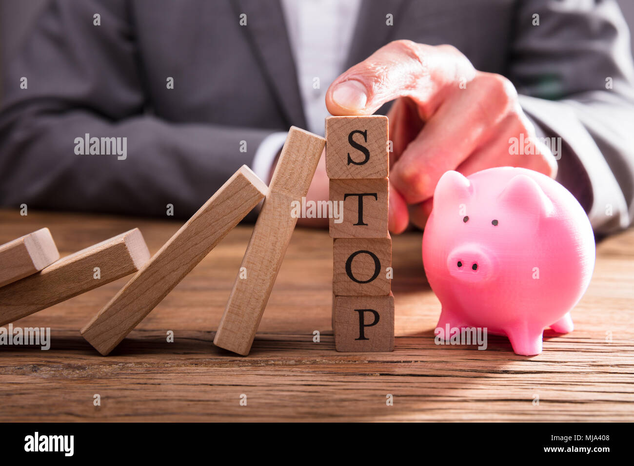 Person's Finger On Stop Wooden Blocks Stopping Dominos From Falling Over Piggybank Stock Photo