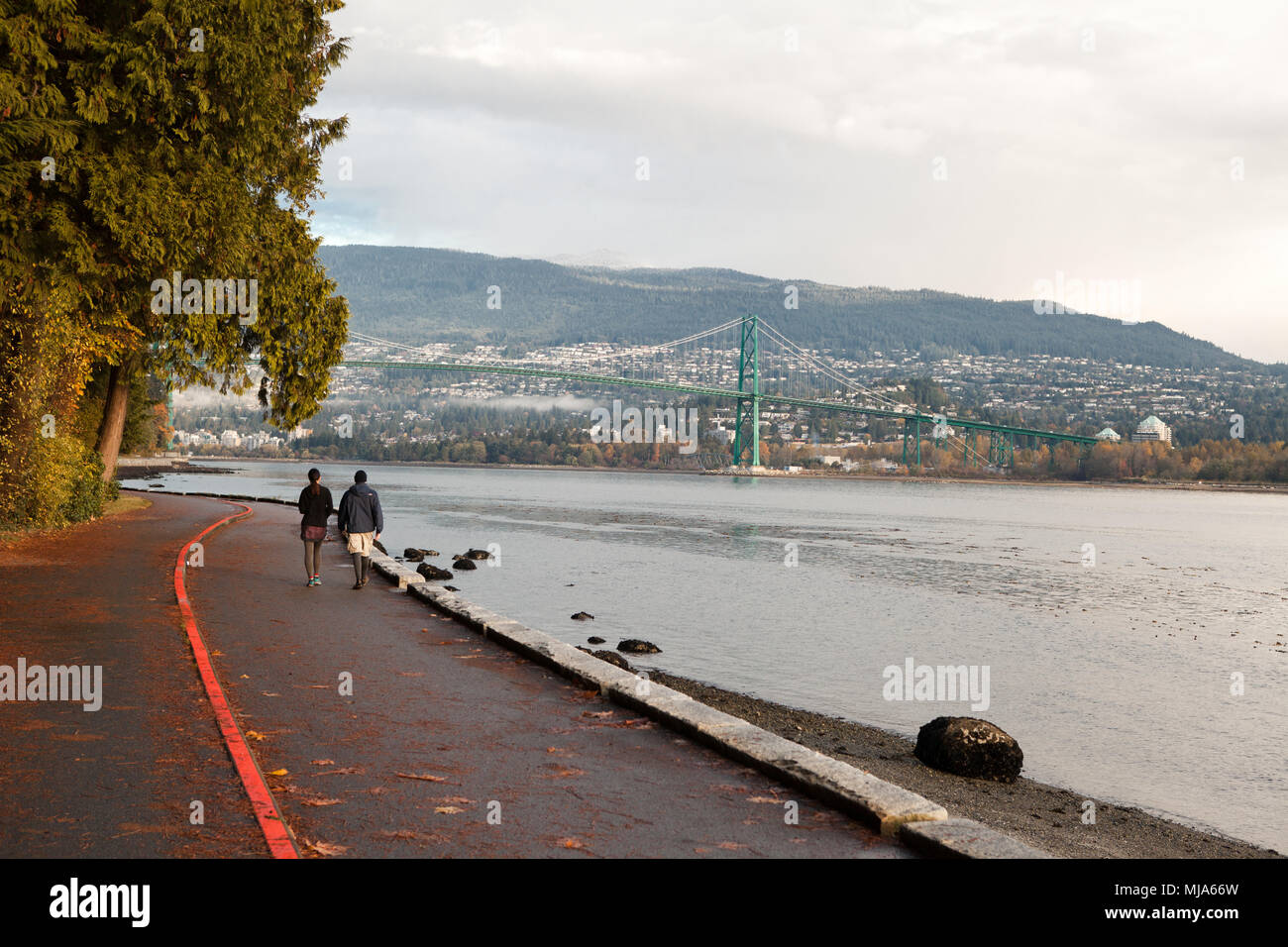 Couple walking on Stanley Park Seawall during the fall season. The golden bridge and north vancouver in the background. Stock Photo