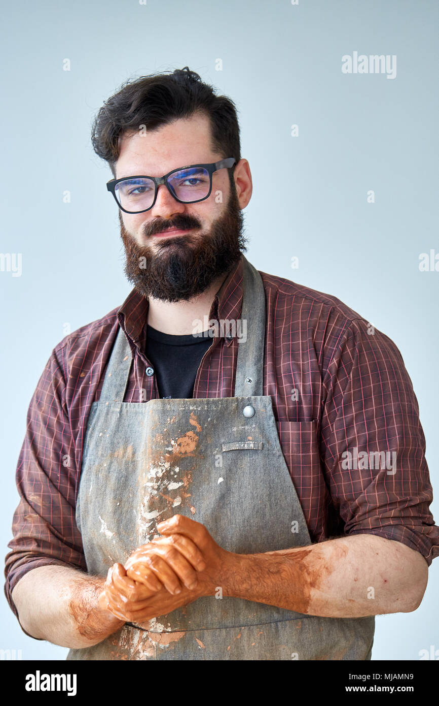 Portrait of handsome young bearded potter posing in studio, close-up. Artistic craftsman wearing plaid shirt and dirty apron holding his hands covered Stock Photo
