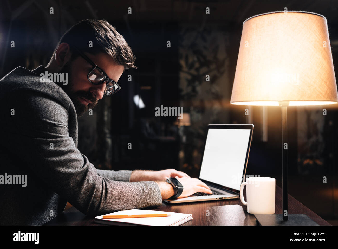 Bearded young coworker wearing glasses typing text on contemporary laptop in modern loft office at night. Business man sits by the wooden table with l Stock Photo