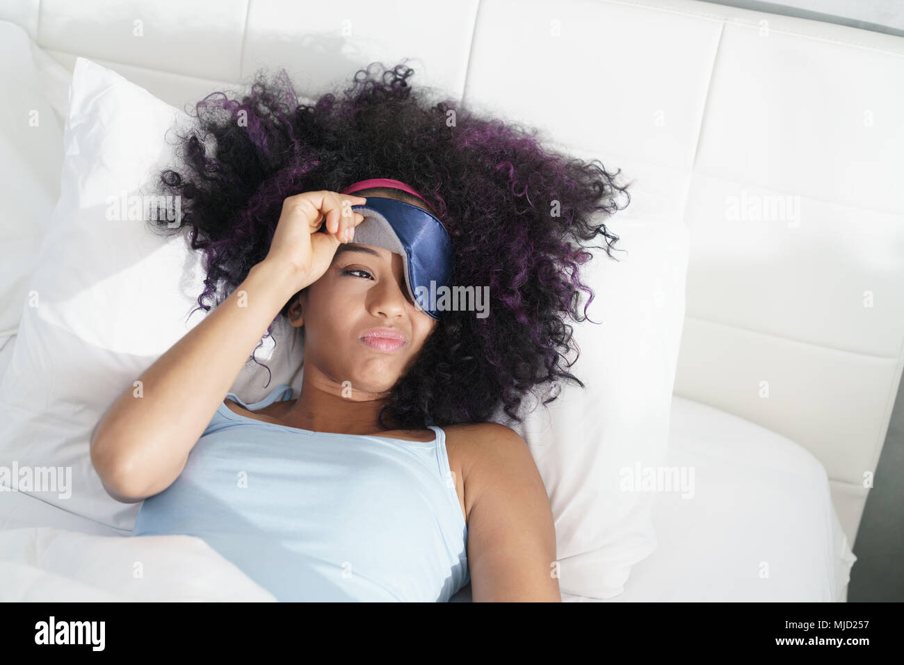 Lazy black girl with blindfold in bed late in the morning. Tired African American woman with sleeping mask sleeping during daytime. Stock Photo