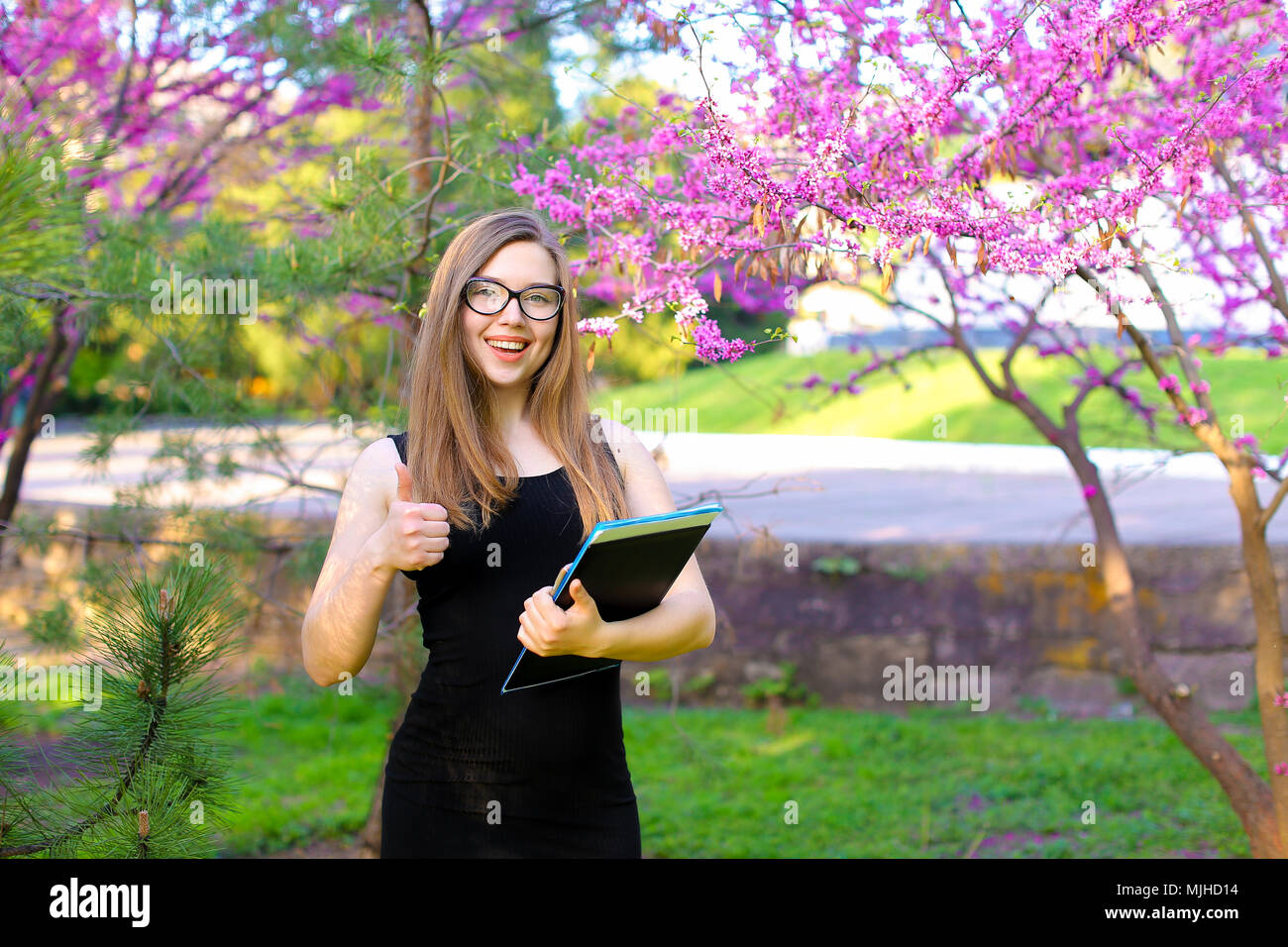 Female student showing thumbs up in garden with documents in black ...