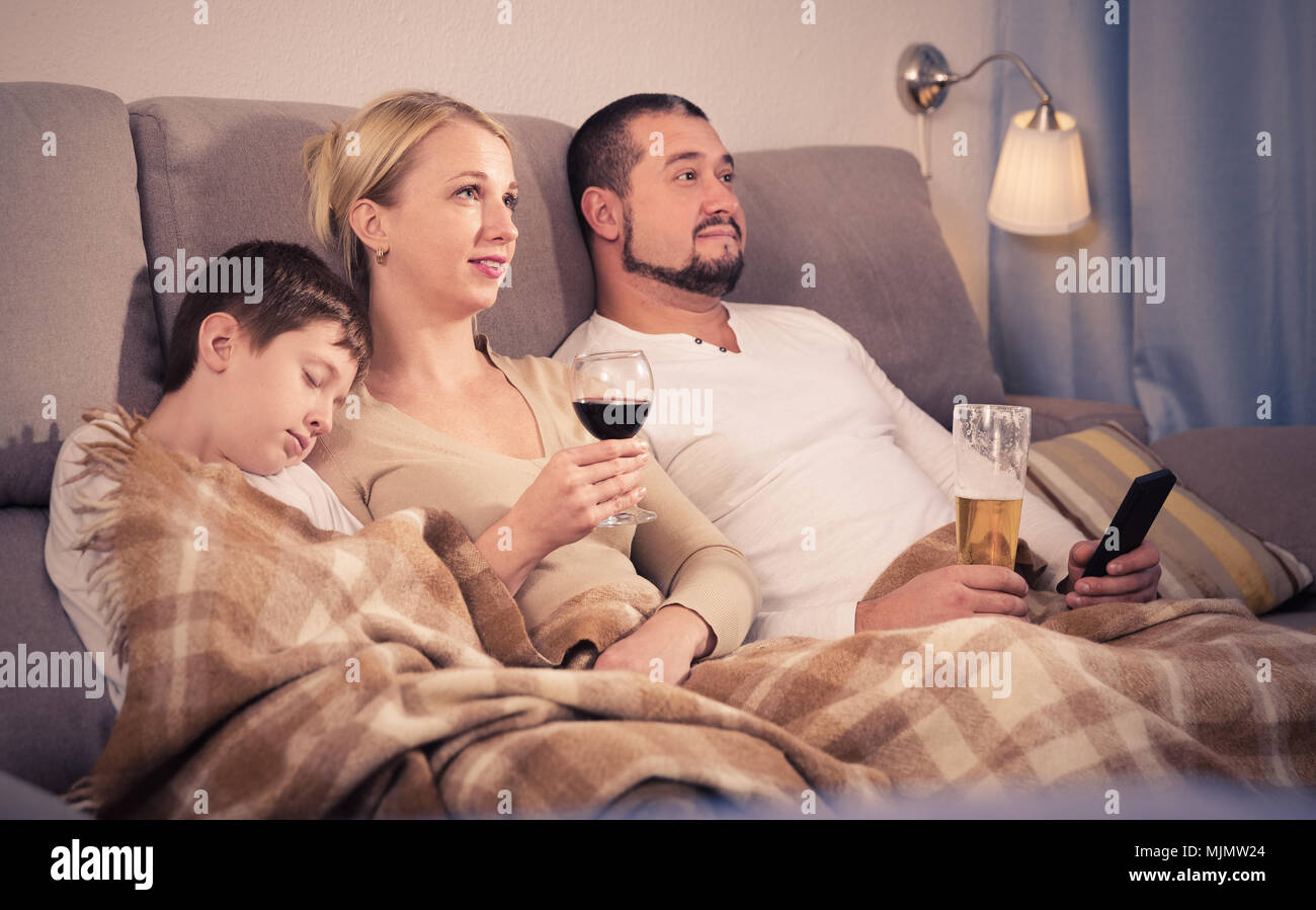 Happy parents lying on sofa under blanket watching TV while son
