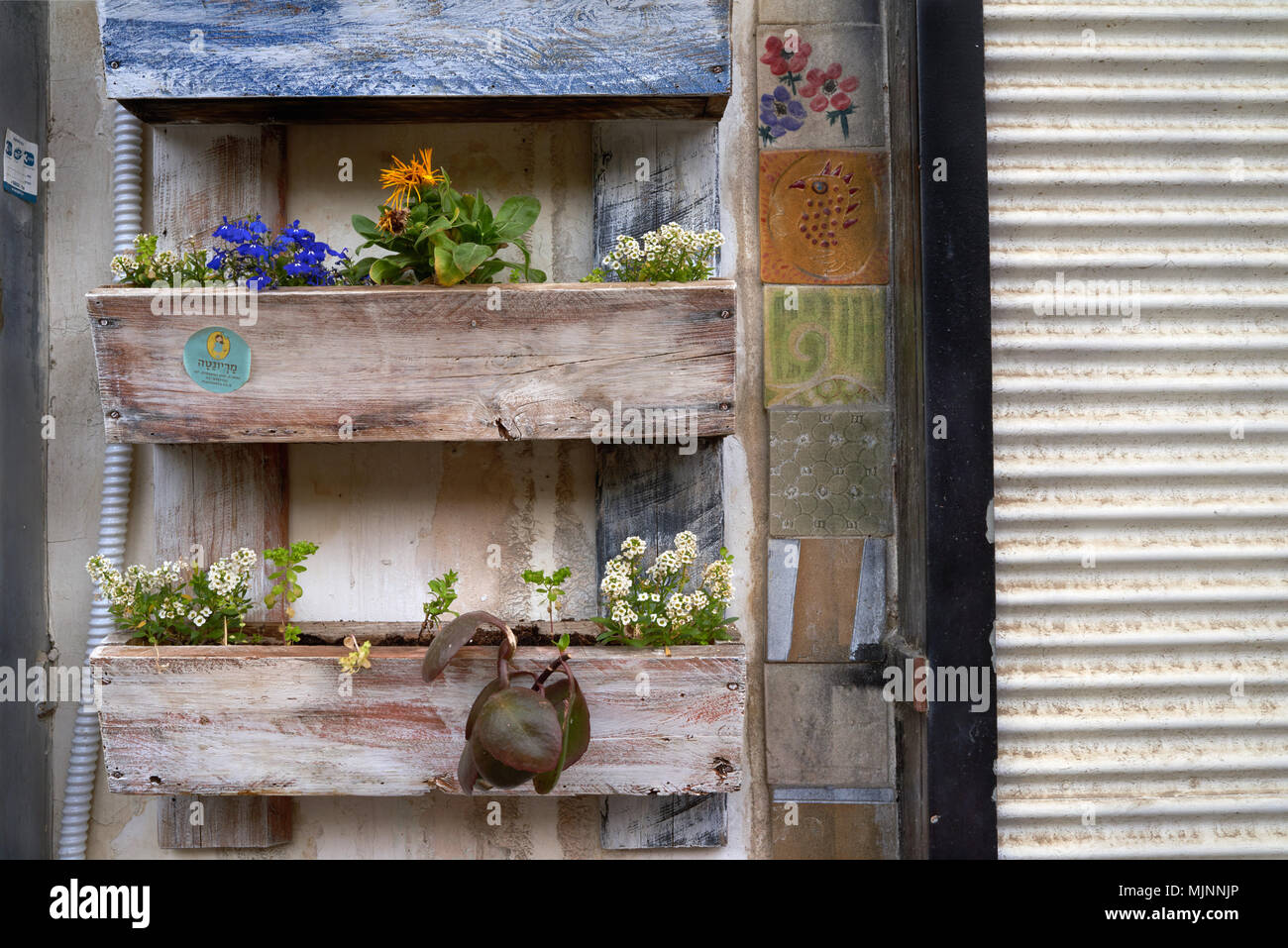 Wooden rectangular boxes with orange flowers on the wall of the house, decorated with multicolored tiles, to the right white roller shutters, urban ba Stock Photo