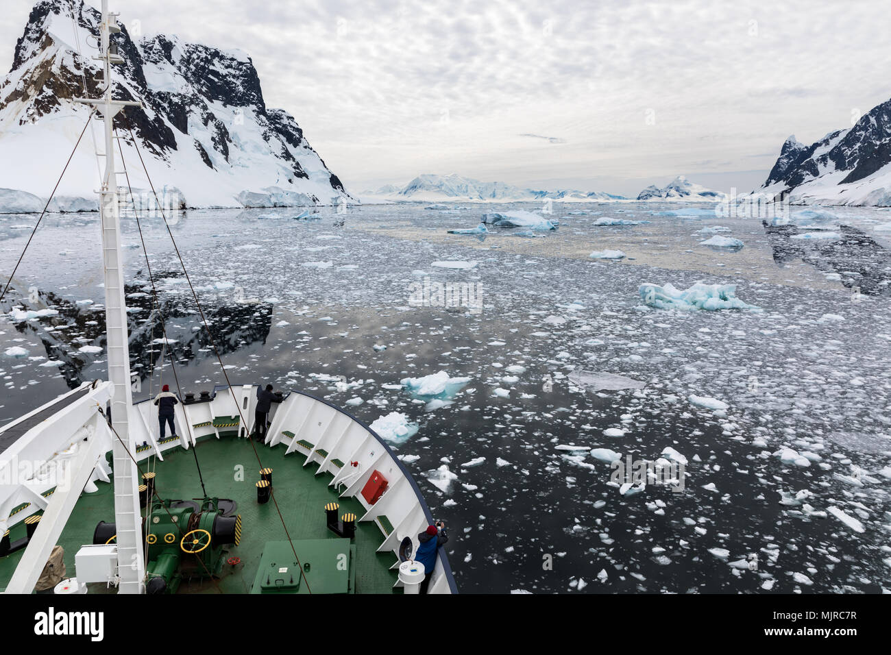 A ship travels through snow covered mountains and water filled with ice while passengers take photos and enjoy the view, Lemaire Channel, Antarctica Stock Photo