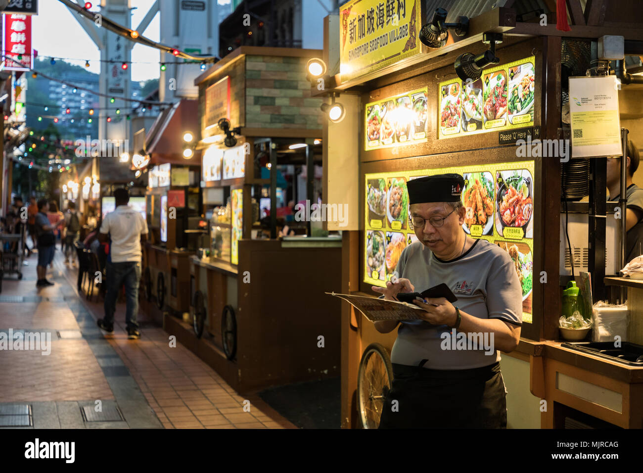 Singapore - March 24, 2018: A food vendor studies his mobile phone while holding a menu at Chinatown's food street as he waits for the evening crowd t Stock Photo