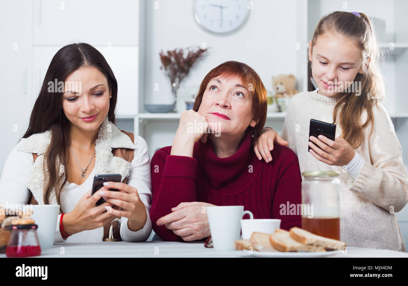 Bored grandma sitting at home table while daughter and granddaughter