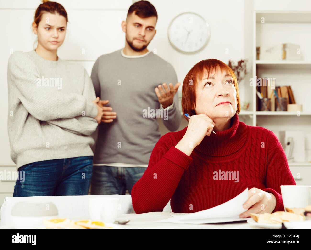 Portrait of distressed aged woman with paperwork at home table with irritated family behind her Stock Photo
