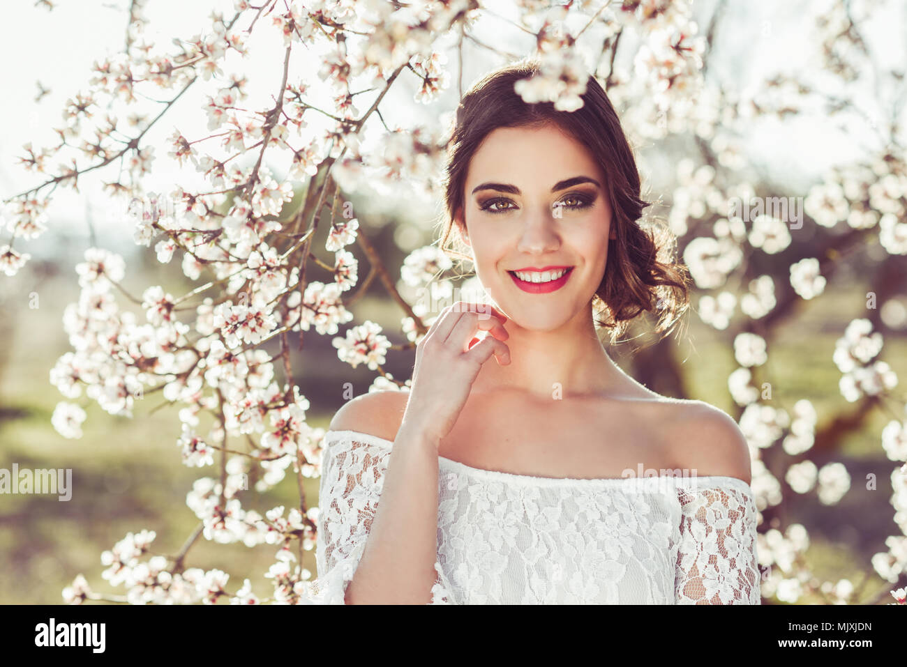 Portrait of young woman smiling in the flowered garden in the spring time. Almond flowers blossoms. Girl dressed in white like a bride. Stock Photo