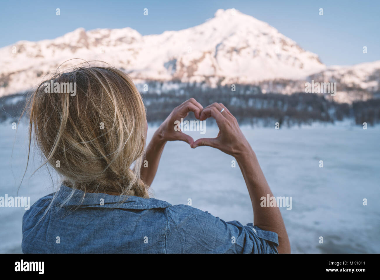 Young woman making a heart shape finger frame over frozen lake and snowcapped mountains. People love nature environment concept. Shot in Switzerland Stock Photo