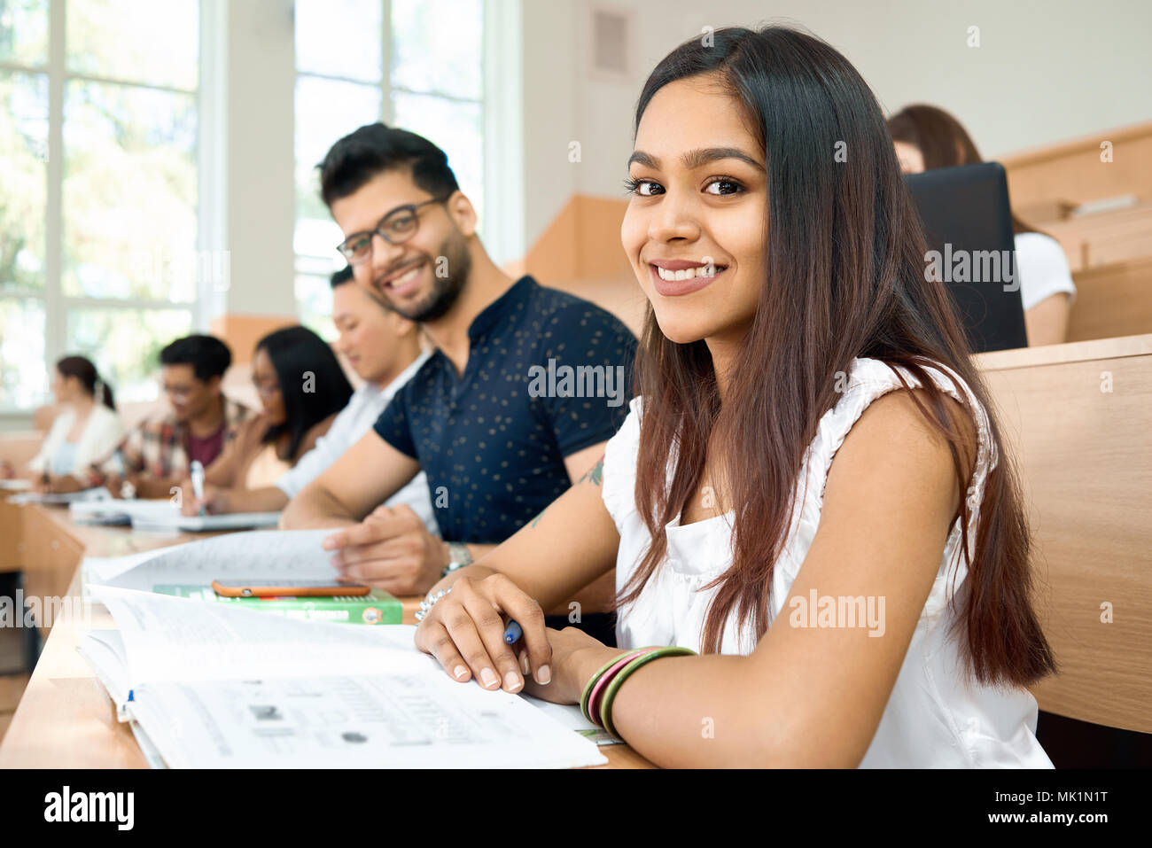 Sideview of students prepearing for exams in university.Girl and boy sitting on wooden desk and looking at camerain spacy modern university classroom with big panoramic windows. Working with project. Stock Photo